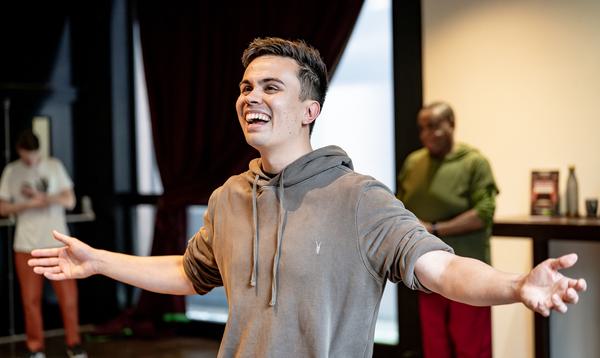 Rishi Manuel is wearing a grey jumper and smiling while looking straight ahead with his arms stretched out on either side of him.