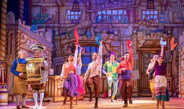 The Dick Whittington and his Cat Ensemble and Max Mirza as Idle Jack are dancing with one hand in the air holding a bit of cloth. Tony Timberlake as Alderman Fitzwarren and Clive Rowe as Sarah the Cook are stood next to them, with Clive Rowe wearing a gold money till.