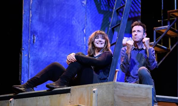 A photo of Josie Lawrence and Lee Simpson sat down together on stage during An Improbable Musical. Josie is smiling and Lee looks angry.