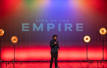 A photo of Nish Kumar on stage during Live at the Empire. Nish is wearing all black and the words LIVE AT THE EMPIRE are displayed on the wall behind him.
