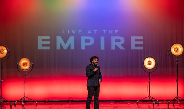 A photo of Nish Kumar on stage during Live at the Empire. Nish is wearing all black and the words LIVE AT THE EMPIRE are displayed on the wall behind him.