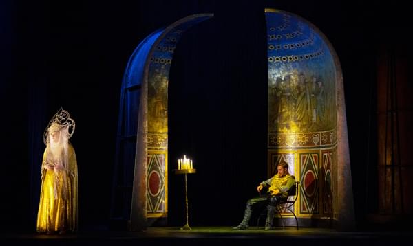 A photo of English Touring Opera's Ottone in performance. A man is sat on stage in a chair whilst a woman with a large headdress faces away from him.