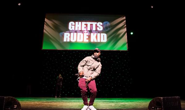 A photo of Ghetts on stage at Hackney Empire, wearing a jacket and leaning to the left, holding a microphone in his right hand.