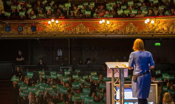 A photo taken from the back of Hackney Empire's stage showing a woman talking to a crowd as they hold up green signs that say TRUE