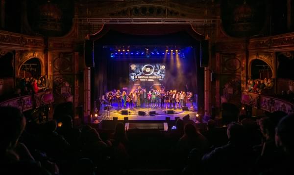 A photo taken from the back of the Dress Circle in Hackney Empire showing a full audience watching all of the people involved in the UK Americana Award 2023 stood together on stage.