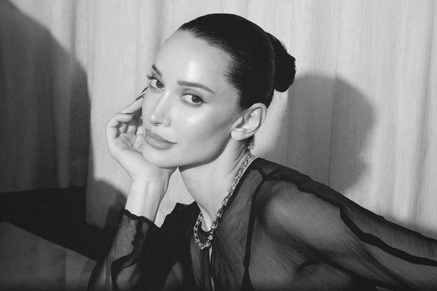 A black-and-white portrait of Roxie with her hair pulled into a sleek bun, resting her head lightly on one hand. She gazes toward the camera with a calm, poised expression and wears a dark, sheer long-sleeved top with a layered chain necklace. Soft, directional lighting creates dramatic shadows against a light curtain backdrop.