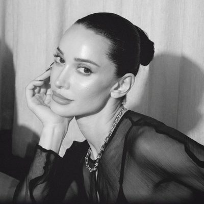 A black-and-white portrait of Roxie with her hair pulled into a sleek bun, resting her head lightly on one hand. She gazes toward the camera with a calm, poised expression and wears a dark, sheer long-sleeved top with a layered chain necklace. Soft, directional lighting creates dramatic shadows against a light curtain backdrop.