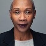 Josette Bushell-Mingo wearing a black blazer and grey t-shirt and staring straight at camera with purple lipstick