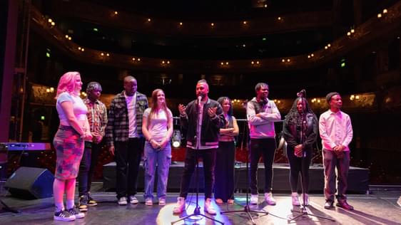 A group of young people stand alongside Yamin Choudury (Artistic Director of Hackney Empire) on Hackney Empire's main stage, preparing to perform.