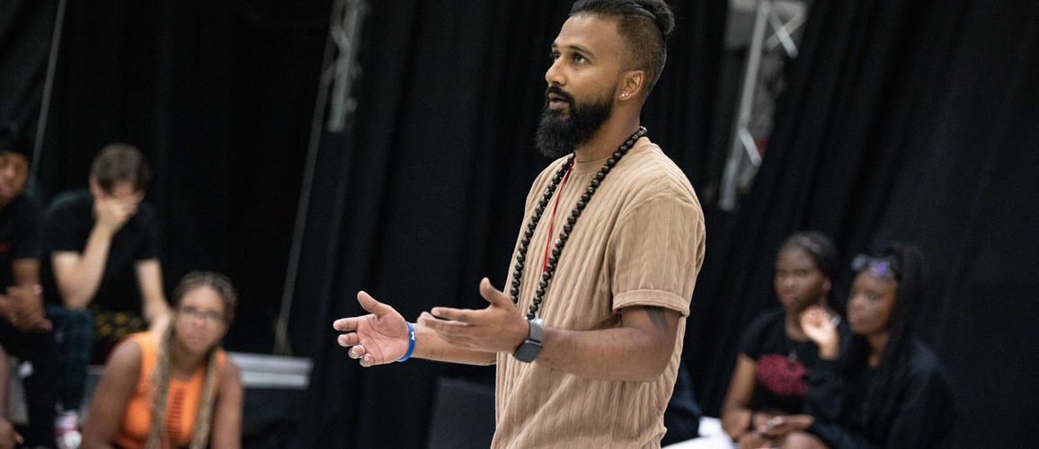 Yamin Choudury directing a group of young people, wearing a brown t-shirt with his arms outstretched.