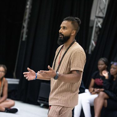 Yamin Choudury directing a group of young people, wearing a brown t-shirt with his arms outstretched.