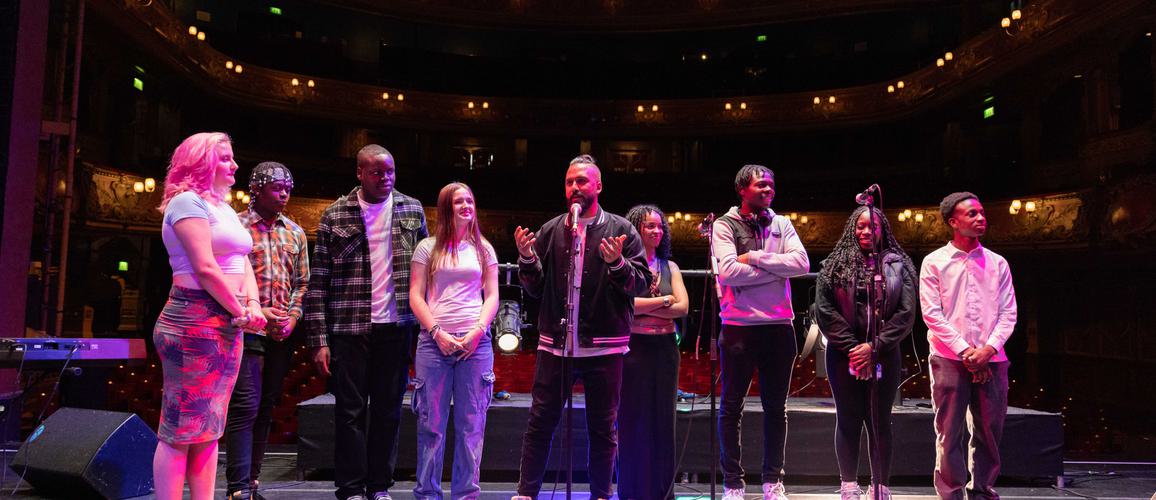 A group of young people stand alongside Yamin Choudury (Artistic Director of Hackney Empire) on Hackney Empire's main stage, preparing to perform.