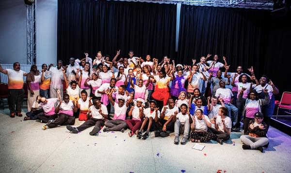 A photo in Hackney Empire's rehearsal space showing over 50 young people in colourful pink, purple and orange t-shirts posing together, many with their arms in the air.