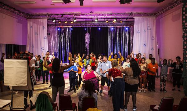 A photo taken from the back of the rehearsal space showing a group of young people in colourful t-shirts performing together.