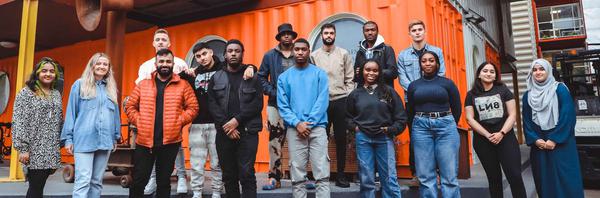 16 participants and organisers for Compulsory Subjects lined up together against an orange background