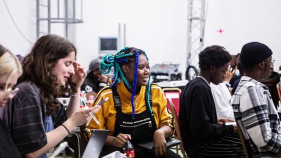 A Creative Futures workshop focusing on a young woman wearing a yellow top and black dungarees with bright blue braids. She is sat surrounded by other young people.