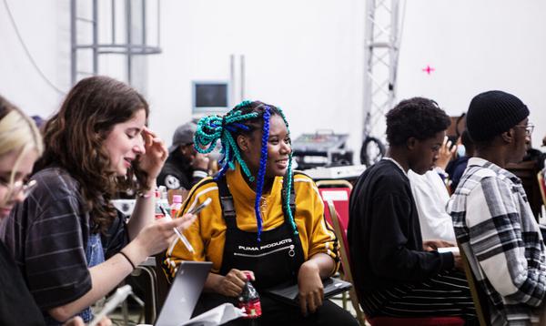 A Creative Futures workshop focusing on a young woman wearing a yellow top and black dungarees with bright blue braids. She is sat surrounded by other young people.