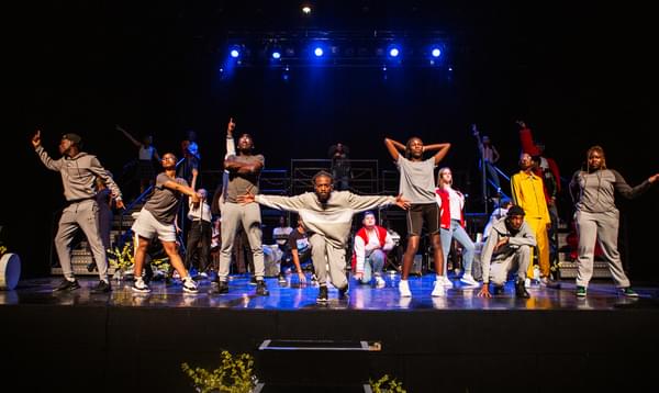 A group of young performers pose along the front of the Hackney Empire stage. Some are standing with arms outstretched, some are kneeling on the ground, but all are doing different poses.