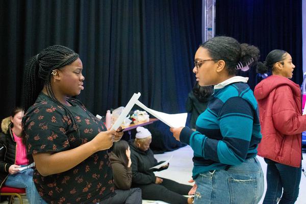 Two young people are holding scripts and facing each other practising some lines. One is wearing a printed black t-shirt and one is wearing a stripey blue top and has glasses.