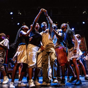 A group of young performers from our Artist Development Programme show: A Direct Message in 2022, stand together in the centre of a stage, crowded round looking at a phone.