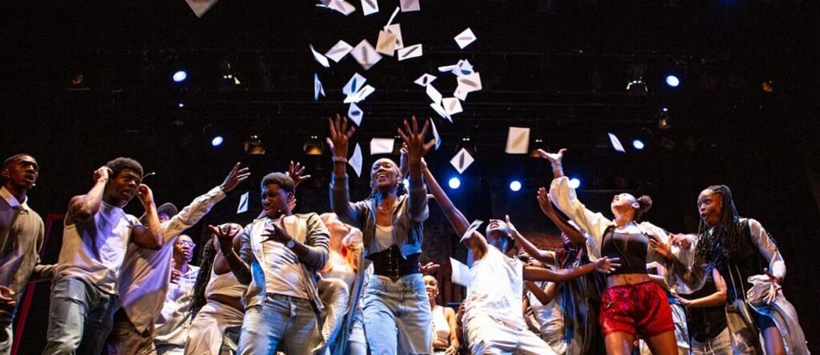 A group of young people crowd in the middle of the stage as pieces of paper are thrown up into the air