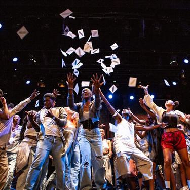 A group of young people crowd in the middle of the stage as pieces of paper are thrown up into the air