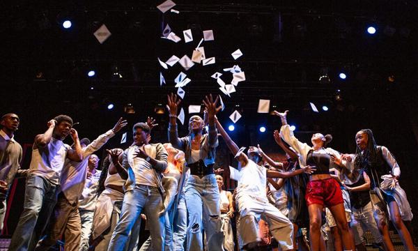 A group of young people crowd in the middle of the stage as pieces of paper are thrown up into the air