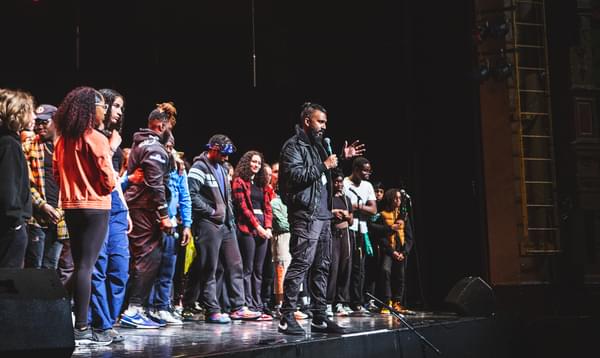 A group of young performers on stage at the end of the show. Artistic Director Yamin Choudury stands at the front of the stage with a microphone talking to the audience.
