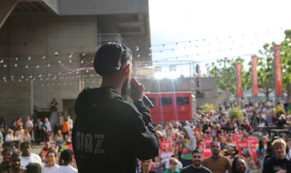 A photo taken from behind a performer at River Stage 2023 showing a crowd on the Southbank watching them. They are wearing a black hoodie.