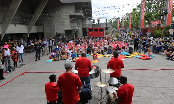 A photo taken from the stage at River Stage 2023 on the Southbank showing the back of some steel pans and drums performers in front of a crowd of people. They are all wearing red t-shirts