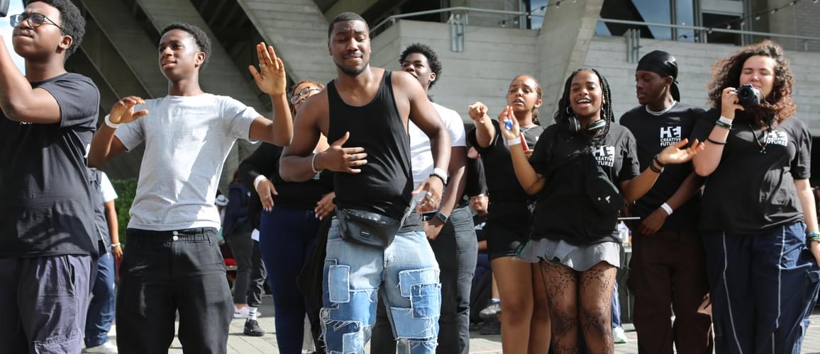 A group of young people are dancing as they watch performers at River Stage 2023 on the Southbank. Many of them are dressed in Hackney Empire t-shirts.