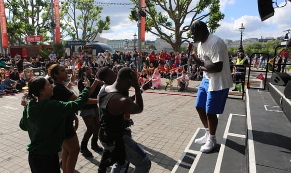 A rapper is performing on the stage at River Stage 2023 on the Southbank. They are wearing a white t-shirt and blue short and performing to a crowd of young people dressed all in black.