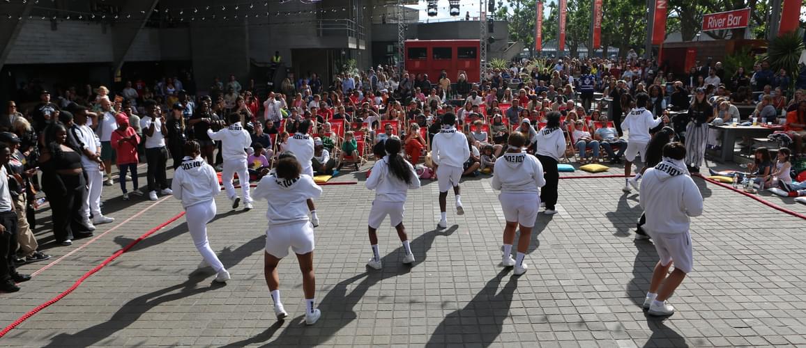 A group of dancers performing at the River Stage on the Southbank in front of a crowd of people. They are wearing white tracksuits and dancing in the sunshine.