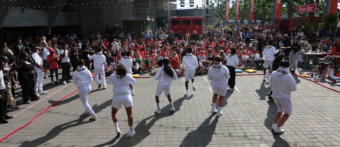 A group of dancers performing at the River Stage on the Southbank in front of a crowd of people. They are wearing white tracksuits and dancing in the sunshine.