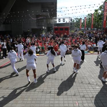A group of dancers performing at the River Stage on the Southbank in front of a crowd of people. They are wearing white tracksuits and dancing in the sunshine.