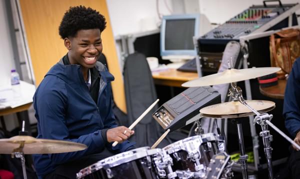 A photo taken during rehearsals for our Artist Development Programme showing a young drummer playing on the drums wearing a blue jacket and smiling.