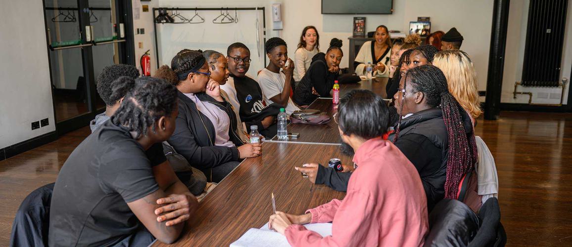 A group of young people are sat opposite each other along a long table discussing ideas. Leona Lewis is sat at the head of the table leading the discussion.