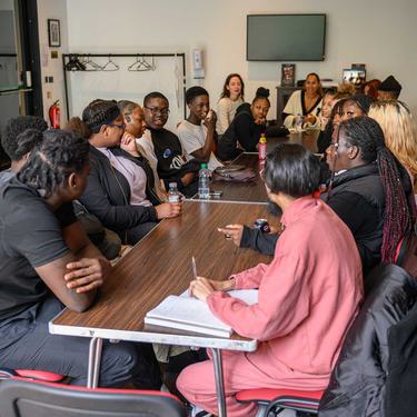 A group of young people are sat opposite each other along a long table discussing ideas. Leona Lewis is sat at the head of the table leading the discussion.