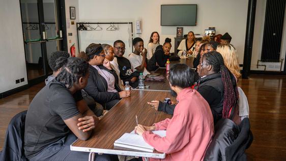 A group of young people are sat opposite each other along a long table discussing ideas. Leona Lewis is sat at the head of the table leading the discussion.