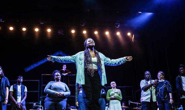 One young person wearing a blue shirt with their arms outstretched is smiling and singing whilst others watch on in the background.