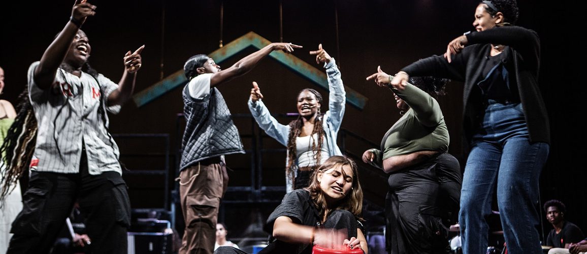 One young person is sat down on the floor drumming onto a bucket whilst six others around them dance and move in response.