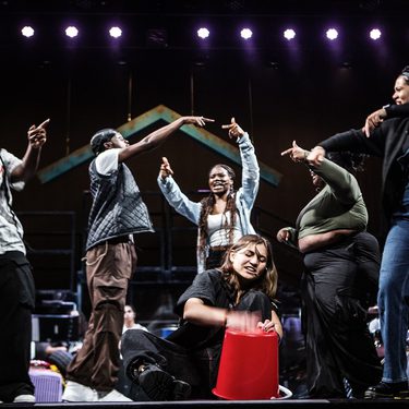 One young person is sat down on the floor drumming onto a bucket whilst six others around them dance and move in response.