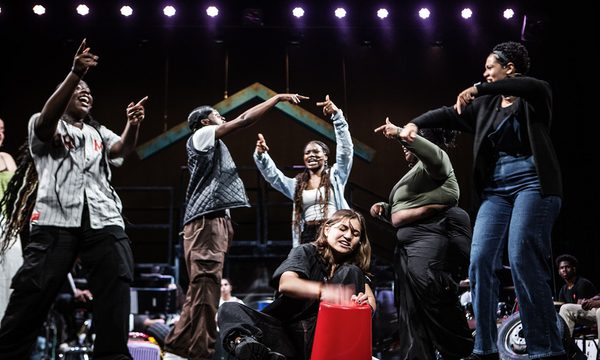 One young person is sat down on the floor drumming onto a bucket whilst six others around them dance and move in response.