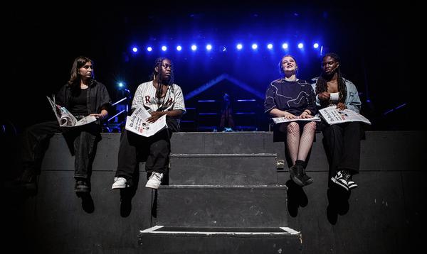 Four young people holding newspapers sit along the front of the stage talking to each other with their legs dangling off.