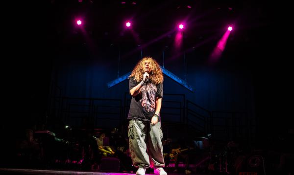 One young person in the centre of the stage wearing a black t-shirt and cargo pants and singing into a microphone.