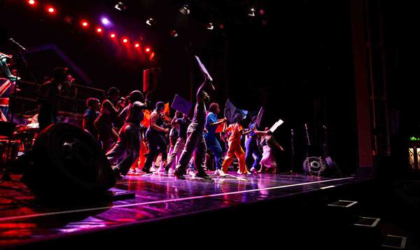 A photo taken of the stage showing a group of young people in colourful clothing all holding up placards in revolution.