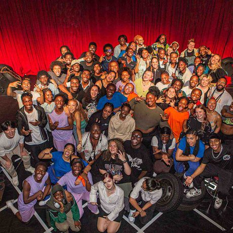 A group shot of the whole Company and Creative Team showing about 70 people posing together on our stage with the red curtain behind.
