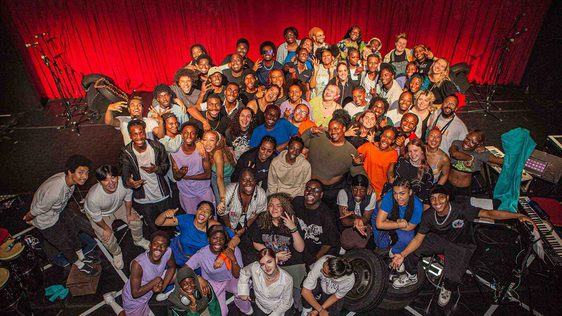 A group shot of the whole Company and Creative Team showing about 70 people posing together on our stage with the red curtain behind.