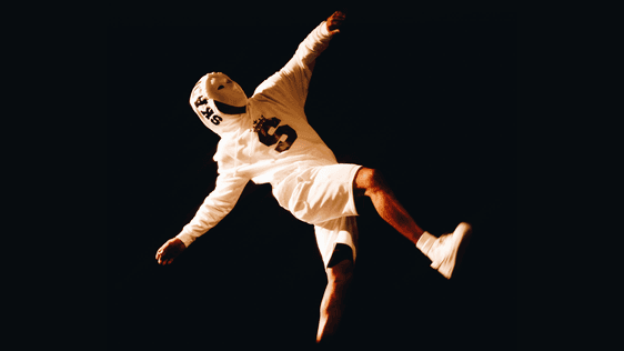 A photo showing one figure wearing a white tracksuit and a white mask in mid air, halfway through a backflip, against a black background.