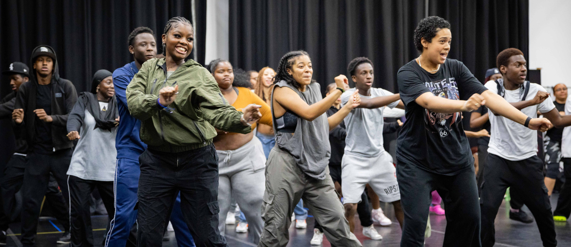 A photo of a group of young people in a rehearsal space warming up by dancing.